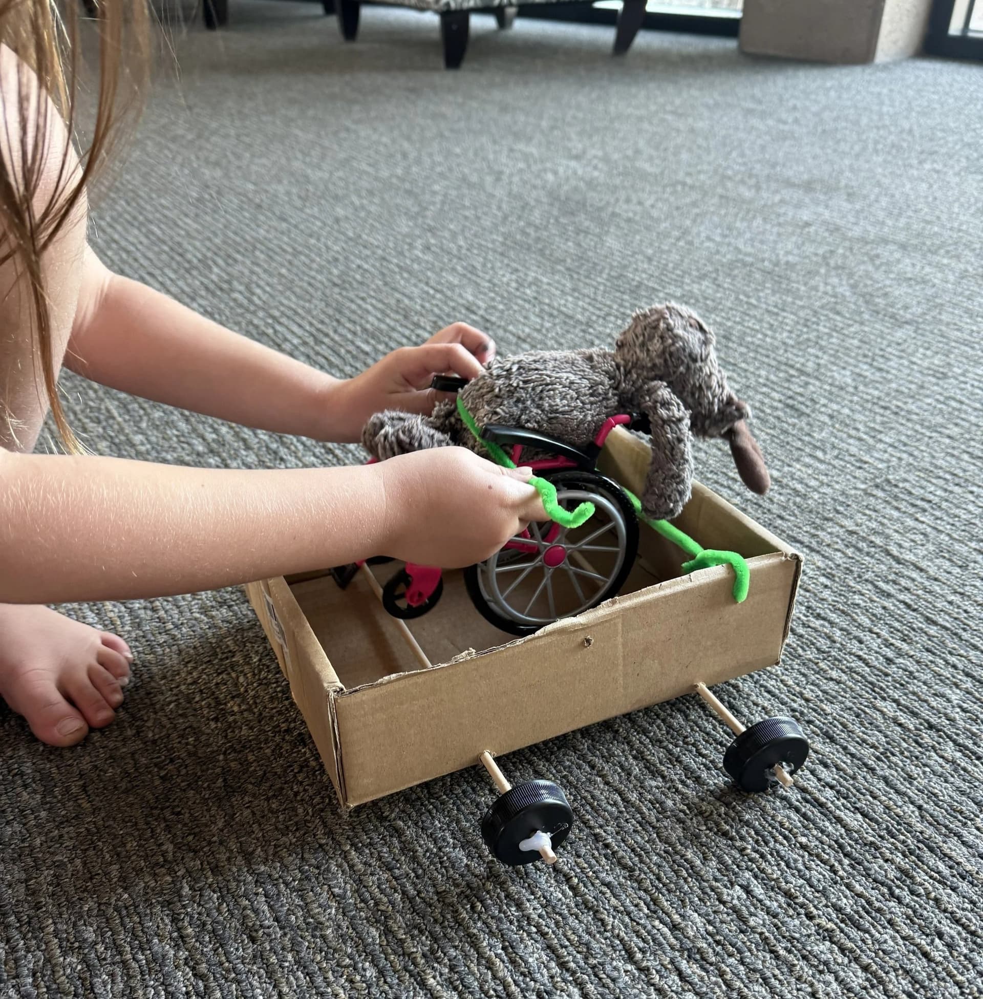 A student-built cardboard vehicle carrying a stuffed animal in a wheelchair